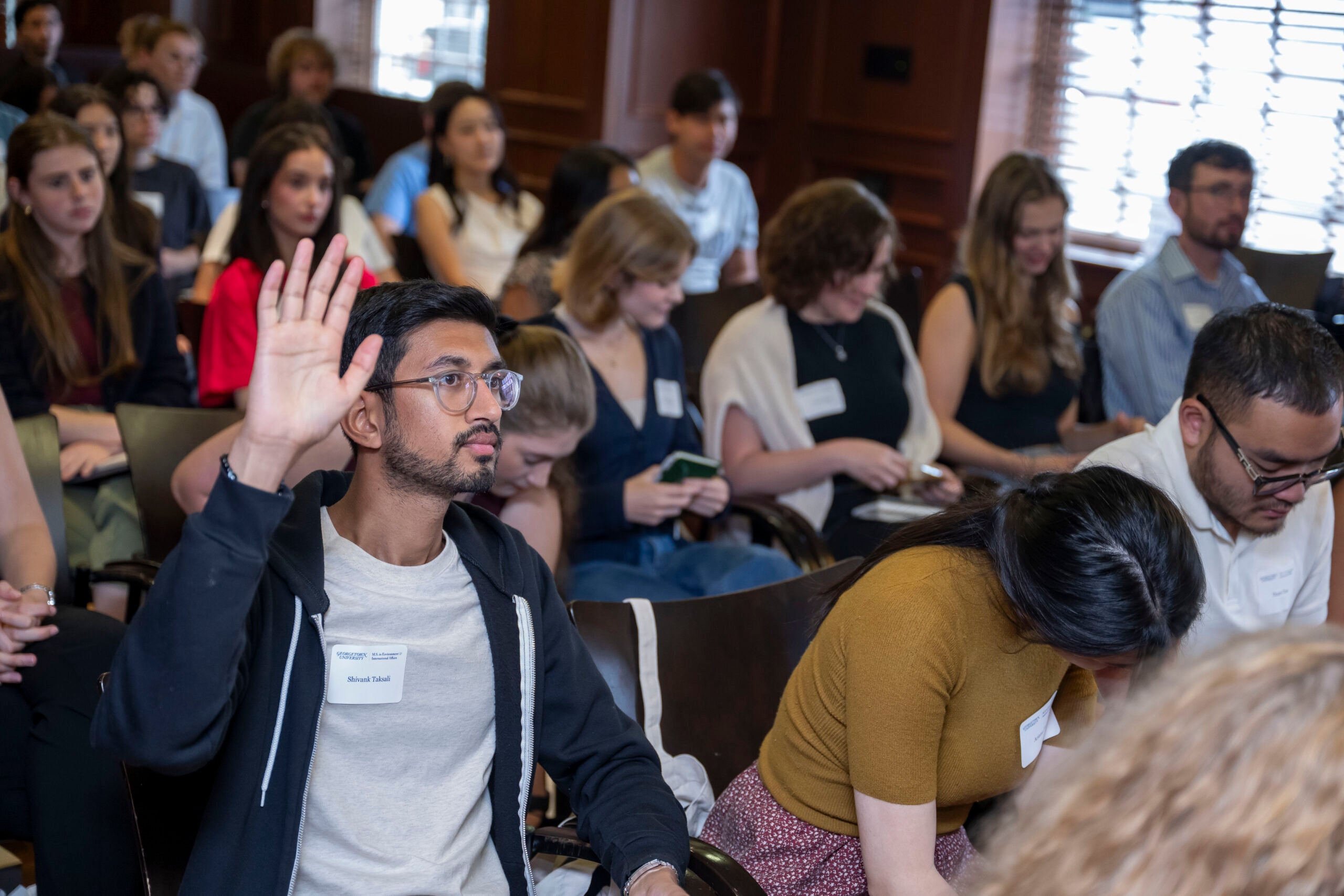 Shivank Taksali sits in a wood-paneled classroom during MS-EIA orientation, raising his hand to participate in discussion. He wears glasses and a Georgetown name tag, while fellow students sit around him listening and taking notes.